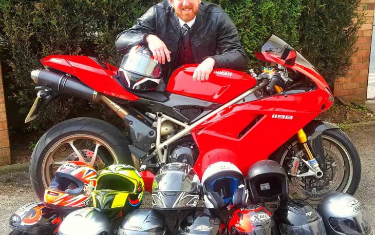 Funeral director leaning on red motor cycle. In front there are 12 helmets.