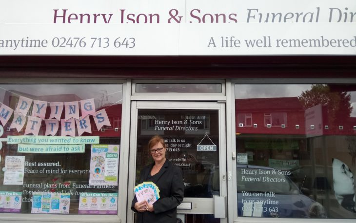 Picture of women standing in front of Henry Ison & Sons shop window. She is holding up a leaflet advertising Dying Matters.