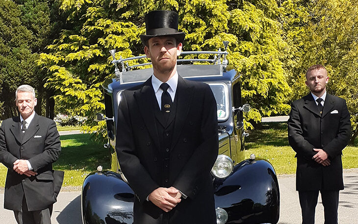 A person in a suit and top hat standing in front of a hearse.