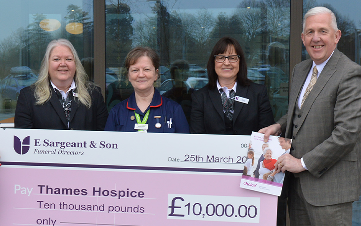 4 people stand in front of glass window holding a large pink cheque raised for £10,000.00.