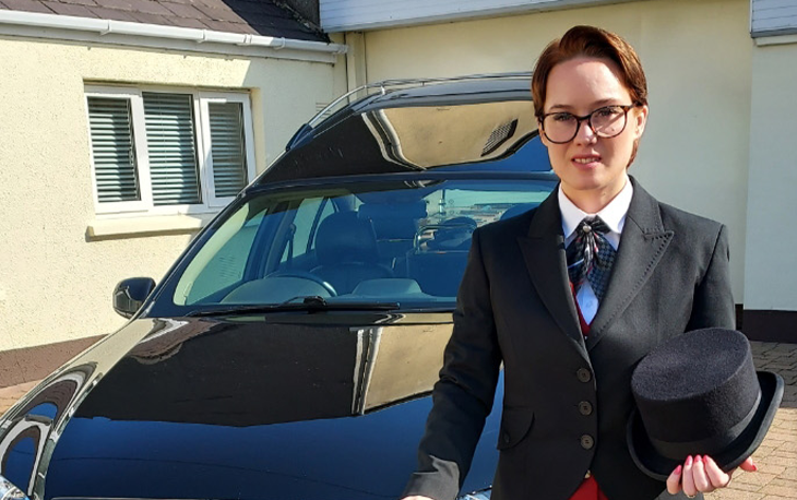 A women in a suit and tie and holding a top hat standing in front of a hearse.