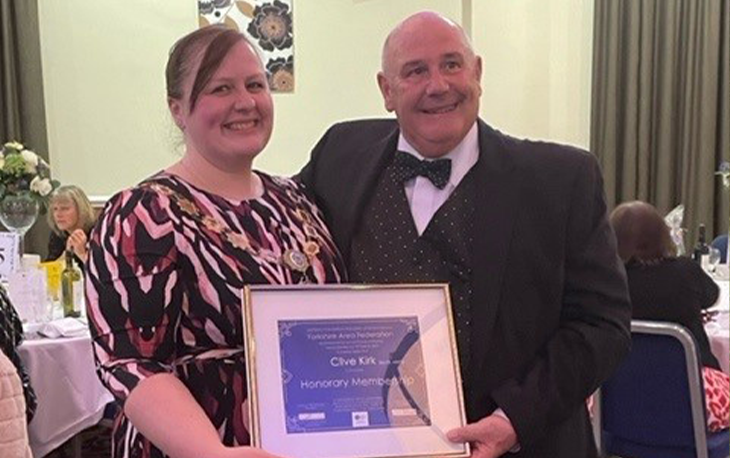 A women in as dress and a man in a suit and bow tie holding up an honorary membership award plaque.