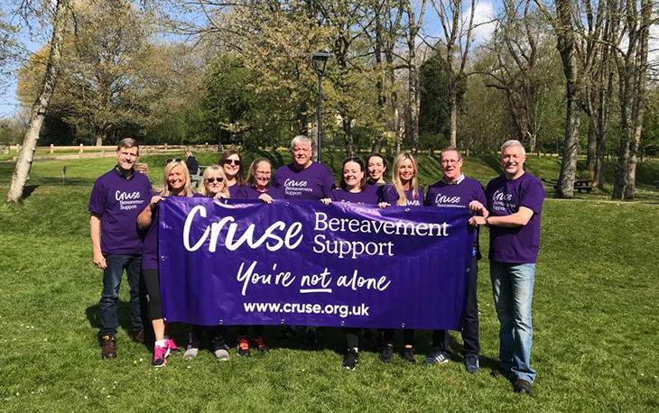 Funeral Director and colleagues stand behind large purple sign which reads Cruse Bereavement Support, you’re not alone.