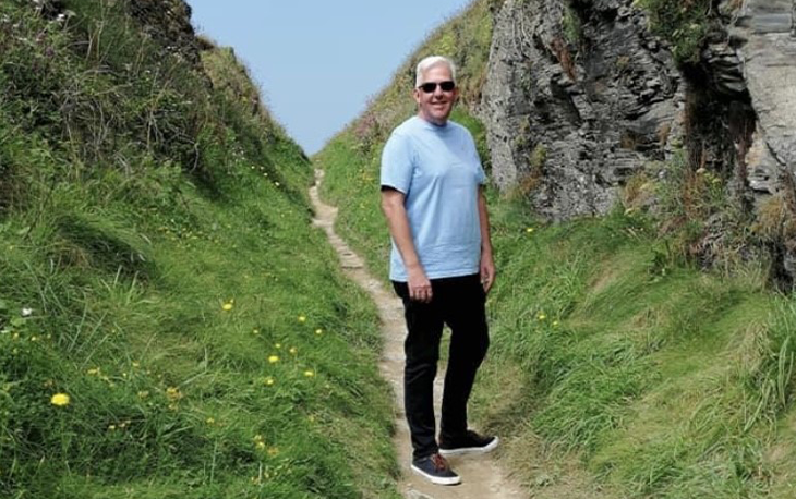 Man hiking on a sandy path between a rock wall and grass hill.