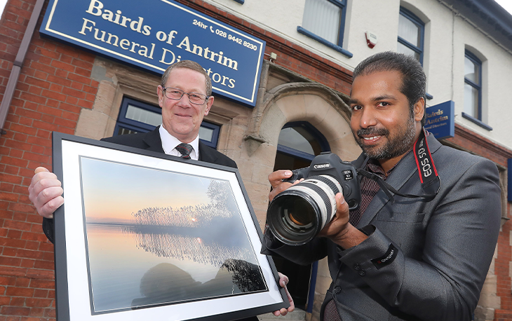 Picture of photographer doling camera and funeral director holding framed picture in front of Bairds of Antrim Funeral Directors.