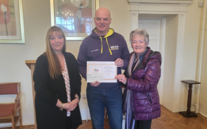 3 people standing in a line, the person in the middle is holding a cheque from fund raising.