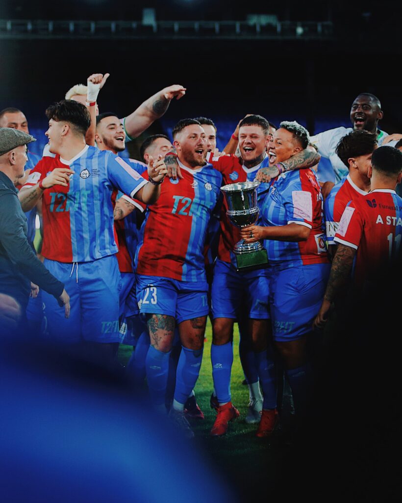 Lianne Sanderson, Jamie Groves, Harry Clark and Dan Spragg huddle around in a semi circle celebrating holding the winners cup. 